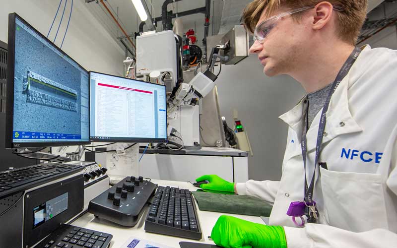 A man sat looking at a computer screen wearing a lab coat and rubber gloves.